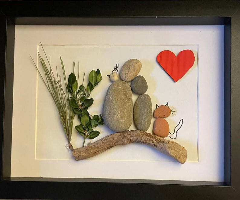 K&ouml;nnte beinhalten: Gerahmtes Kunstwerk mit einer Familienszene aus nat&uuml;rlichen Materialien. Glatte Steine bilden Figuren, ein rotes Papierherz und eine Katze. Gr&uuml;nes Laub und Treibholz verleihen der Komposition Textur.