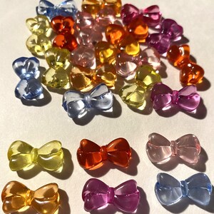 May include: A collection of translucent bow-shaped beads in various colors, including yellow, orange, pink, purple, and blue. The beads are scattered across a white surface, creating a colorful and playful display. These beads are ideal for crafting and jewelry making.