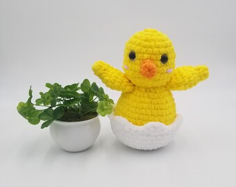 Puede incluir: Un juguete de pollito de crochet amarillo sentado en una cáscara de huevo de crochet blanca. El pollito tiene ojos negros, un pico naranja y mejillas rosadas.