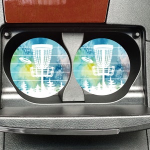 May include: Two round car coasters with a tie-dye design in shades of blue, green, and yellow. The coasters feature a white silhouette of a disc golf basket with a disc flying through the air.