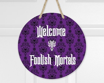 May include: A round purple hanging sign featuring a black damask pattern and the white text Welcome Foolish Mortals with a central white icon. The sign is suspended by a tan rope loop at the top, creating a decorative wall display piece.