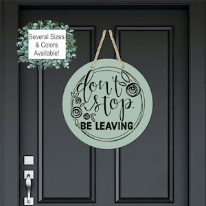 May include: A round, light green wooden sign with black text that reads "Don't Stop Be Leaving." The sign is decorated with black floral designs and is hanging on a dark gray door.