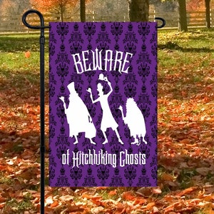 Op de afbeelding: Paarse tuin vlag met een zwart-wit patroon. De vlag heeft een wit silhouet van drie geesten met de tekst "Beware of Hitchhiking Ghosts".