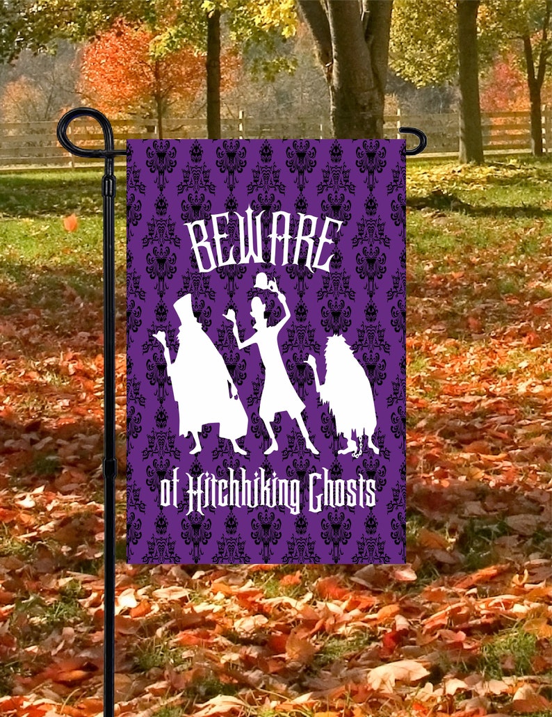 Op de afbeelding: Paarse tuin vlag met een zwart-wit patroon. De vlag heeft een wit silhouet van drie geesten met de tekst "Beware of Hitchhiking Ghosts".