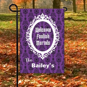 May include: A rectangular garden flag featuring a purple background with a black damask pattern. A white ornate oval frame contains the text Welcome Foolish Mortals in a gothic font. Below the frame, the text The Bailey's is printed in white.