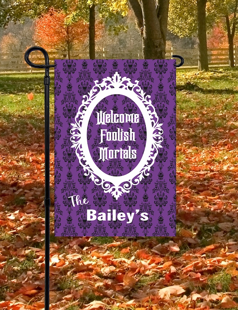 May include: A rectangular garden flag featuring a purple background with a black damask pattern. A white ornate oval frame contains the text Welcome Foolish Mortals in a gothic font. Below the frame, the text The Bailey's is printed in white.