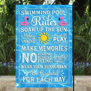 May include: A blue and white swimming pool rules sign with pink flamingos, a yellow sun, and the text "Swimming Pool Rules, Soak Up The Sun, Relax and Unwind, Laugh and Play, Make Memories, No Running Pushing Peeing, Hang Your Wet Towels, Wear Your Sunscreen, Be Grateful For Each Day".