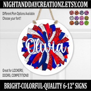 May include: A white round sign with a red, white, and blue pom-pom design. The sign has the name "Olivia" written in a white cursive font. The sign is 6-12 inches in diameter.