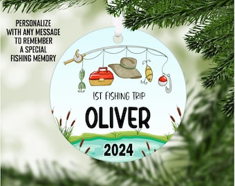 May include: A blue and white round Christmas ornament with a fishing rod, a red tackle box, a hat, and a fish. The ornament says "1st Fishing Trip Oliver 2024".