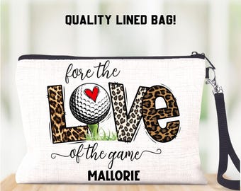 Op de afbeelding: Witte canvas toilettas met een zwarte rits en een zwarte band. De tas heeft een golfbal met een rood hart erop en de woorden "fore the LOVE of the game MALLORIE" in zwarte tekst.