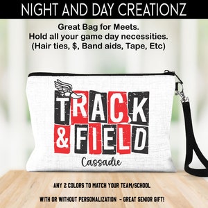 May include: White zippered makeup bag with a black strap. The bag has a red and black graphic that says "TRACK & FIELD" with a running shoe graphic. The name "Cassadie" is written below the graphic. The bag is on a wooden surface.