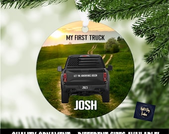 Op de afbeelding: Een ronde kerstbal met een zwarte pick-up truck met een Amerikaanse vlag op de achterruit. De truck rijdt over een onverharde weg met een zonsondergang op de achtergrond. De tekst "My First Truck" staat bovenaan de kerstbal, en de tekst "Let the Adventures Begin" staat op de achterkant van de truck. Het jaar "2023" en de naam "Josh" staan ook op de kerstbal.