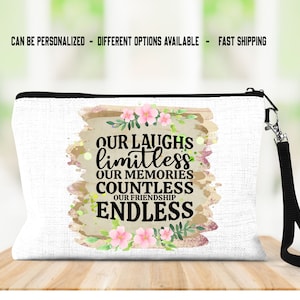 May include: A white canvas makeup bag with a black zipper and a black strap. The bag has a floral design with pink flowers and green leaves. The text on the bag reads "Our laughs limitless, our memories countless, our friendship endless."