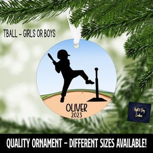 May include: Round ornament featuring a black silhouette of a baseball player swinging a bat. The ornament has a blue sky background and a baseball field. The name "OLIVER" and the year "2023" are printed on the ornament. The text "TBALL - GIRLS OR BOYS" is at the top.