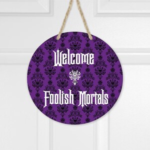 May include: A circular purple hanging sign featuring a black damask pattern with hidden skull motifs. The text "Welcome" and "Foolish Mortals" is printed in white in a gothic font, with a white decorative emblem centered between the two lines.