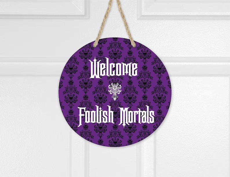 May include: A circular purple hanging sign featuring a black damask pattern with hidden skull motifs. The text "Welcome" and "Foolish Mortals" is printed in white in a gothic font, with a white decorative emblem centered between the two lines.