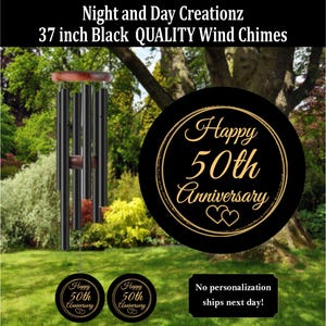 May include: Black wind chimes, 37 inches long, with a wooden top and accents. The chimes are displayed outdoors with a black circular sign that reads "Happy 50th Anniversary". Additional small signs with the same text are also visible.