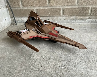 May include: A brown and red spaceship model with a pointed nose and two long wings. The spaceship has a white symbol on the side.