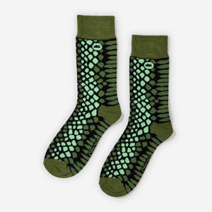 May include: A pair of crew socks featuring a snake scale design in green and black. The socks have a dark green cuff, heel, and toe. A small logo is visible on the cuff. The socks are designed to be worn on the feet.
