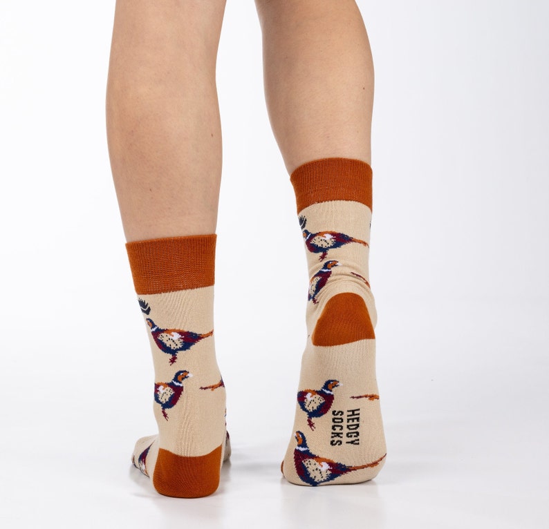 May include: A pair of beige socks with a brown pheasant print. The socks have a brown cuff and the text "HEDGY SOCKS" is printed on the back of one sock.