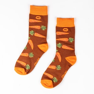 Könnte beinhalten: Ein Paar braune Socken mit orangefarbenem Bund und einem Muster aus orangefarbenen Karotten auf braunem Hintergrund. Die Socken haben das Wort "lucky" seitlich aufgedruckt.