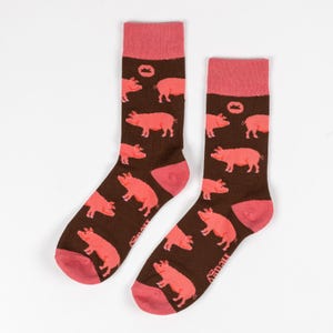 Peut inclure: Une paire de chaussettes marrons avec un motif de cochons roses. Les chaussettes ont un revers rose et un petit logo couronne.