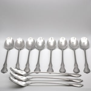 May include: A set of eleven silver-toned metal spoons arranged with eight spoons standing upright in a row and three spoons lying horizontally in the foreground. Each spoon features a polished finish and a decorative embossed pattern on the handle.