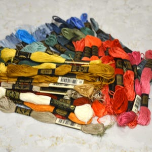 May include: A collection of embroidery floss skeins in a variety of colors including yellow, orange, red, pink, blue, green, and beige. Each skein is secured with a black paper label featuring the DMC brand logo and a barcode for identification.