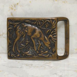 May include: A rectangular bronze-toned belt buckle featuring a raised relief design of a deer standing among foliage. The metal surface has a dark, weathered patina with textured details throughout the scene and a smooth, open loop on the right side.