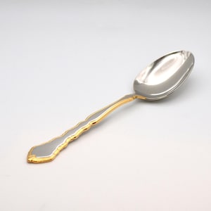 May include: A silver spoon with a gold-colored handle. The spoon's bowl is oval-shaped and highly polished, reflecting the light. The handle has a wavy design with gold trim, adding a touch of elegance to the utensil.