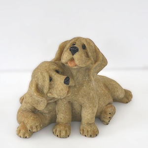 May include: A pair of tan-colored dog figurines. One dog is sitting, and the other is leaning against it. The dogs have black eyes and open mouths, with a light orange tongue visible. The figurines are on a white background.