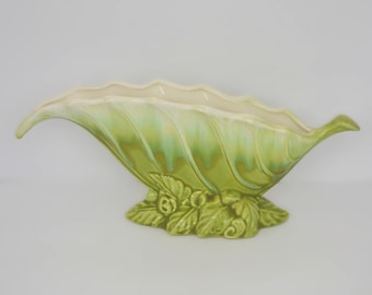 May include: A ceramic leaf-shaped vase in shades of green and cream. The vase has a textured, ribbed design and is supported by a base with leaf and berry details. The interior is cream-colored.