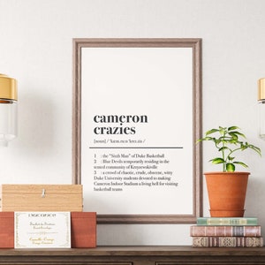 May include: A framed print with a definition of "cameron crazies" in black text on a white background. The print is hanging on a white wall above a wooden console table with two drawers. There are two gold pendant lights hanging above the table, and a potted plant is on the table next to a stack of books.