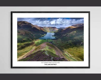 May include: A framed print of Buttermere from Fleetwith Pike in The Lake District. The landscape photograph features a lake surrounded by green hills and mountains under a cloudy sky. The print is in a black frame.