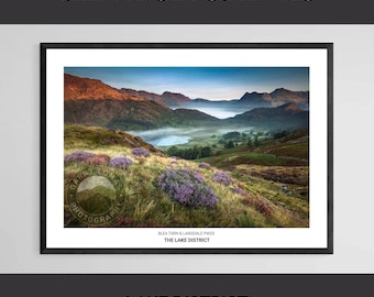 May include: Framed landscape print featuring a scenic view of the Lake District. The image showcases a lake surrounded by rolling hills and mountains, with purple flowers in the foreground. Text at the top reads "BLEA TARN & LANGDALE PIKES" and "LAKE DISTRICT" at the bottom.