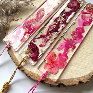 Pu&ograve; includere: Quattro segnalibri rettangolari trasparenti con fiori pressati rosa e rossi e glitter dorato. Ogni segnalibro ha un nome diverso scritto in lettere dorate in corsivo: "Bonnie", "Vanessa", "Levi" e "Emma".