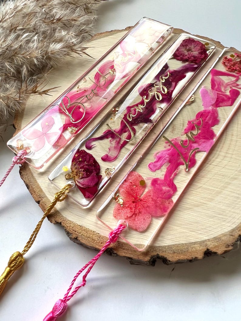Pu&ograve; includere: Quattro segnalibri rettangolari trasparenti con fiori pressati rosa e rossi e glitter dorato. Ogni segnalibro ha un nome diverso scritto in lettere dorate in corsivo: "Bonnie", "Vanessa", "Levi" e "Emma".