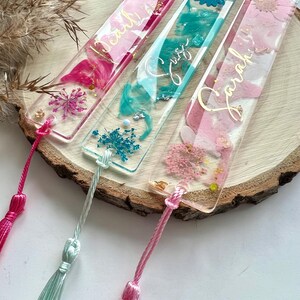 Pu&ograve; includere: Tre segnalibri rettangolari trasparenti con fiori pressati e lettere dorate. I segnalibri hanno fiori rosa, blu e turchese e sono attaccati a nappe rosa, turchese e bianche.