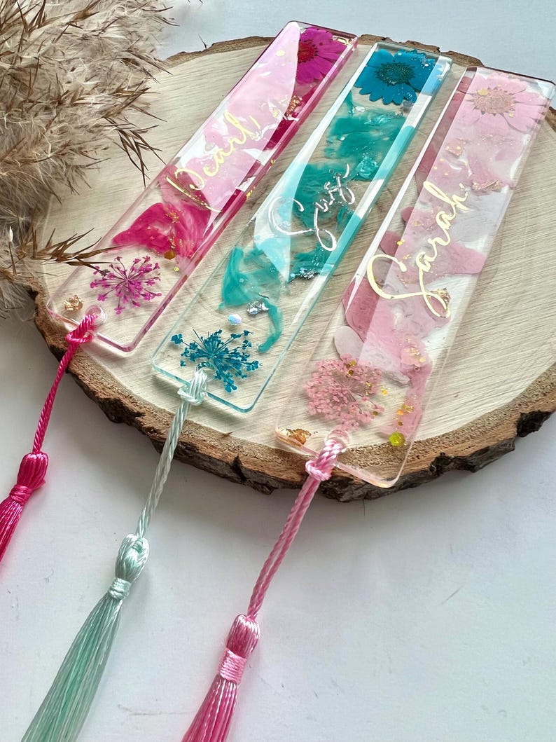 Pu&ograve; includere: Tre segnalibri rettangolari trasparenti con fiori pressati e lettere dorate. I segnalibri hanno fiori rosa, blu e turchese e sono attaccati a nappe rosa, turchese e bianche.