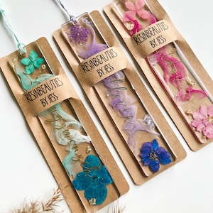 Pu&ograve; includere: Tre segnalibri in resina fatti a mano con fiori pressati e dettagli in foglia d'oro. Ogni segnalibro ha uno schema di colori diverso: turchese, viola e rosa. I segnalibri sono etichettati "Resinbeauties by Jess".