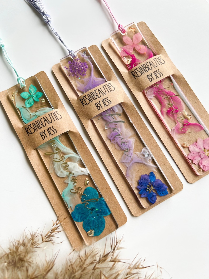 Pu&ograve; includere: Tre segnalibri in resina fatti a mano con fiori pressati e dettagli in foglia d'oro. Ogni segnalibro ha uno schema di colori diverso: turchese, viola e rosa. I segnalibri sono etichettati "Resinbeauties by Jess".