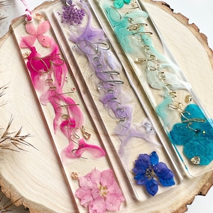 Pu&ograve; includere: Tre segnalibri colorati con fiori pressati e lettere dorate. I segnalibri dicono "Booklover" in corsivo. I segnalibri sono rosa, viola e blu.