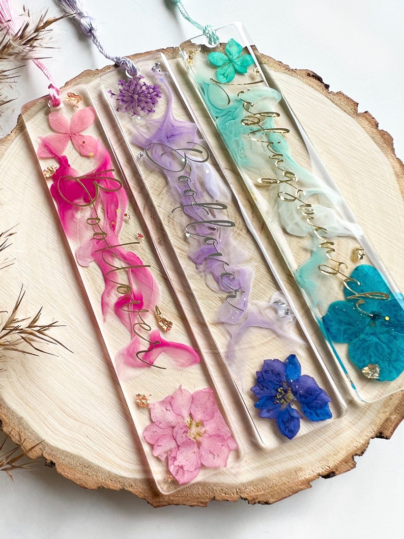 Pu&ograve; includere: Tre segnalibri colorati con fiori pressati e lettere dorate. I segnalibri dicono "Booklover" in corsivo. I segnalibri sono rosa, viola e blu.