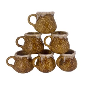 May include: Set of six small, brown ceramic mugs with a textured diamond pattern. Each mug has a handle and a white, dripped glaze around the rim. The mugs are stacked in a pyramid shape against a white background.
