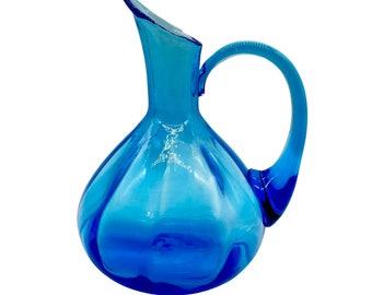 May include: A blue glass pitcher with a wide, rounded body and a curved handle. The pitcher has a textured surface and a slightly iridescent finish.