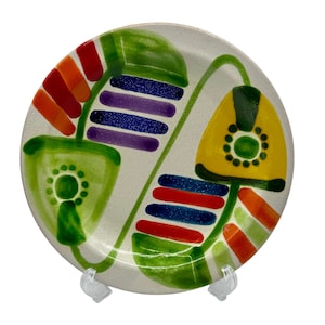 May include: A white ceramic plate with a colorful abstract design. The design features green, yellow, red, orange, blue, and purple shapes. The plate is on a white stand.