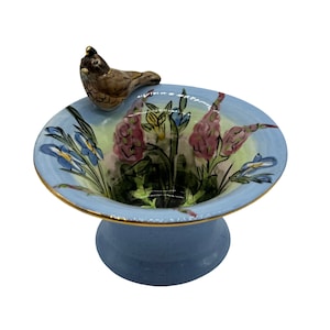 May include: A blue ceramic bird bath with a gold rim and a small bird perched on the edge. The bath is decorated with a floral design featuring pink, blue, and yellow flowers.