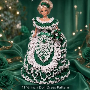 May include: A doll wearing a dark green and white crochet dress with a ruffled off-the-shoulder neckline, a white lace-style apron overlay, and a matching green floral hair accessory. The text reads CROCHET PATTERN Digital Download 11 1/2 inch Doll Dress Pattern.