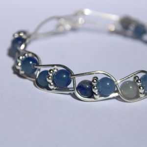 May include: A silver wire bracelet with blue gemstone beads. The bracelet is made of a silver wire that is twisted and woven together to create a unique design. The beads are a mix of light and dark blue shades.