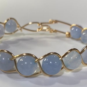 Op de afbeelding: Een delicate gouden draad armband met lichtblauwe edelsteen kralen. De armband is gemaakt met een geweven draad ontwerp en beschikt over een reeks ronde, gladde stenen.